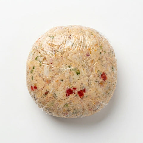 Raw 7 oz Pelican crab cake wrapped in film — chunky crabmeat, red pepper, and herbs on white